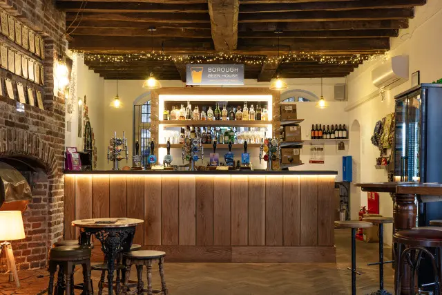 The bar at the Borough Beer House, a clean wood panelled bar with lights underneath the bar counter. above the bar is a beamed ceiling with fairy lights attached to the beams. To the left of the bar is a bare brick fireplace. Behind the bar are illuminated shelves with bottles on them. Two large black fridges are to the right of the bar.
